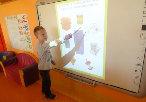 Chłopiec stoi pod tablicą interaktywną i wskaźnikiem pokazuje gazetę.